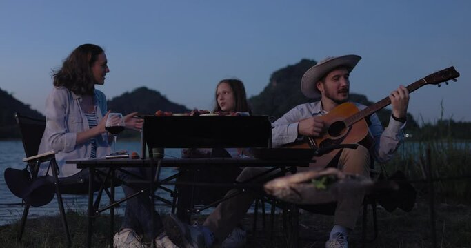 Happy family backpacker eat BBQ and drink and playing guitar together with happiness in Summer while having camping near lake at twilight time, 4k video