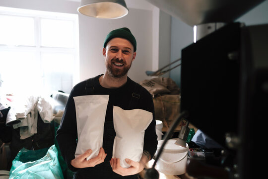 Smiling coffee roaster holding packed paper bags of coffee beans in factory