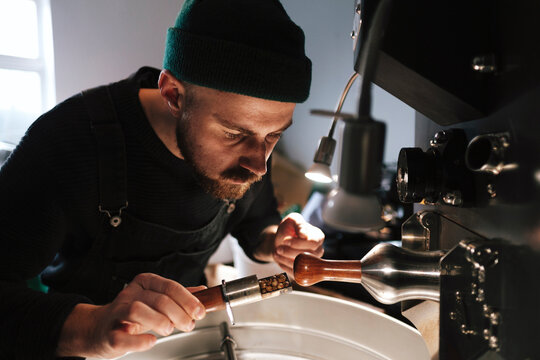 Coffee roaster wearing knit hat and examining coffee beans near machinery