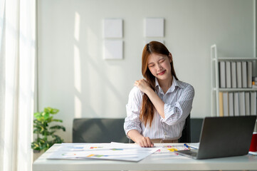 A woman is sitting at a desk with a laptop and a stack of papers. She is wearing a striped shirt and has her arm around her neck. The scene suggests that she is working on a project or task