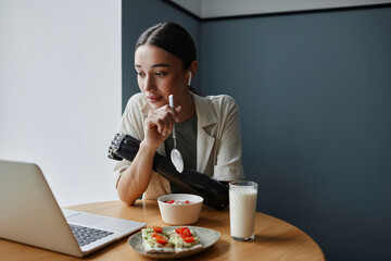 Young woman with bionic arm eating healthy breakfast and watching video on laptop at table