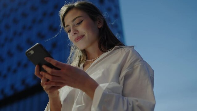 Serene young woman in white blouse looks down at her phone, cityscape backdrop, texting or browsing on a sunny day. Young Professional Woman Using Smartphone in Urban Setting