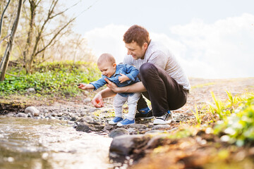 Fototapeta premium Father playing with little toddler boy throwing rocks in water. Boy and dad having fun during warm spring day. Father's day concept.