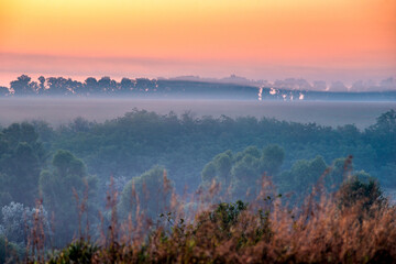 Fototapeta premium Red sunrise over the field with trees and forest . Morning at summer . Fog over the field and trees . Beautiful landsapes . Red sky and blue hour on nature . Smoothly foggy morning 