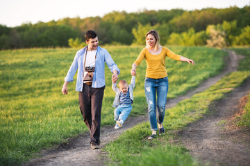 Fototapeta premium New parents holding small toddler, by hands, swinging him. Family outdoors on walk in spring nature. Happy family moment