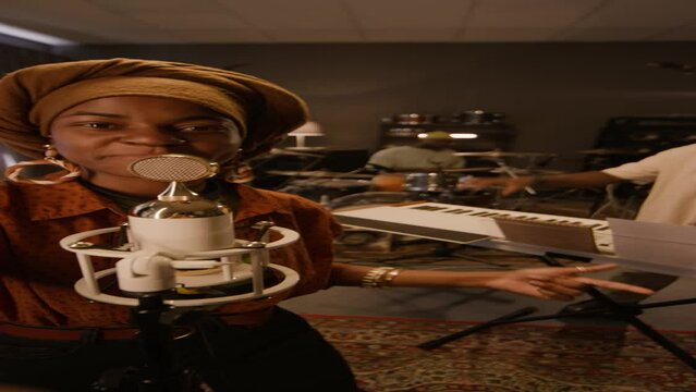 Medium vertical UGC of African American female musician in ethnic turban singing into vintage microphone, filming herself on smartphone for fans on social media, while rehearsing with bandMedium verti
