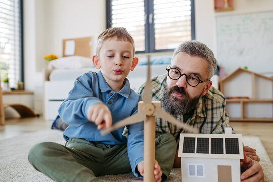 Father explaining renewable green energy, teaching son about sustainable lifestyle. Playing with model of house with solar panels, wind turibine at home. Learning through play.