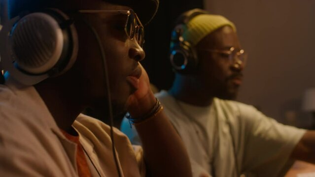 Medium side close-up of two African American electronic musicians sitting in recording studio, listening to new hip-hop music track in headphones, enjoying it and rocking to rhythmMedium side close-up