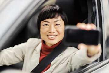 Portrait of a japanese woman sitting in a car and showing cellphone at the camera.