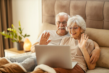 Smiling senior couple sitting on a bed in a bedroom and having video call on a laptop.