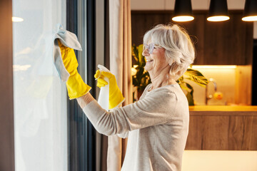 Neat senior woman is cleaning windows at home with detergent and cloth.