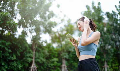 fit, health, exercise, people, outside, looking, relaxation, fitness, sport, lifestyle. A woman is walking in a park while listening to music. She is wearing headphones and holding a smartphone.