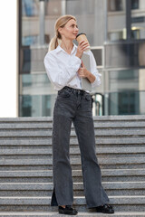 Fototapeta premium Slender young blond business woman dressed in a white shirt carries a cup of coffee and a laptop in her hands while walking