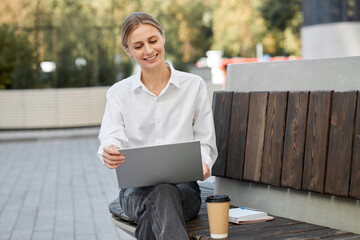 A young blond woman in a white blouse drinks coffee and sits on a bench during a break from work