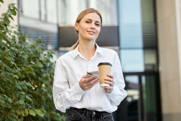 Cute blond woman dressed in a white shirt drinks coffee at lunch while walking on the street