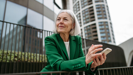 Mature businesswoman holding smartphone, waiting for business partner in the city. Beautiful older woman with gray hair standing on city street.