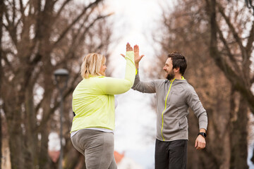 An overweight woman running in nature with friend, high five. Exercising outdoors for people with obesity, support from friend, fitness coach.