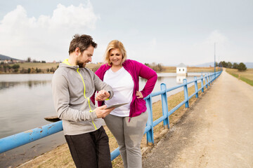 Overweight woman resting after run, personal trainer checking her profile, performance on tablet. Exercising outdoors for people with obesity, support from fitness coach.