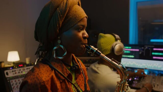 Medium tilting close-up young African American female musician playing saxophone solo, smiling male sound engineer operating mixing console, while recording music for new jazz album in studio