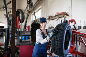 Female auto mechanic changing tieres in auto service. Beautiful woman holding tire in a garage,...