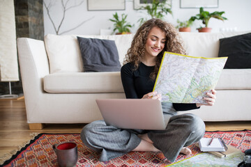 Beautiful woman planning summer vacation abroad, going on trip alone. Sitting on floor, working on itinerary on laptop and looking at map.