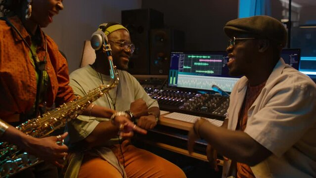Medium of young African American female musician with saxophone, two black male bandmates listening to recording of new track in studio, swaying to rhythm, chatting and laughing