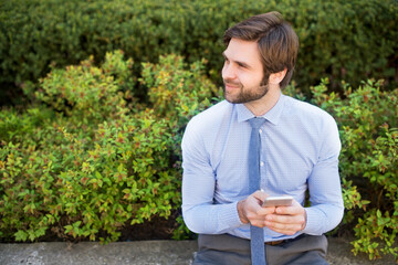 Handsome businessman holding smartphone sitting on bench in city park. Working remotely, waiting for business meeting. Manager smiling, outdoor in urban setting