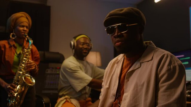Medium of African American male musician in cap and sunglasses playing piano keyboard, woman performing on sax, audio engineer in headphones manning mixing board, while recording music in studio