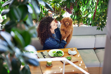Portrait of beautiful curly brunette woman embracing pet while enjoying plant-based meal in a vegan...