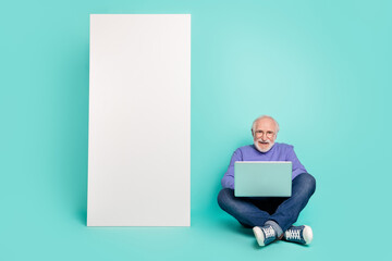 Full length photo of cheerful aged man sit floor use netbook huge empty space telephone isolated on cyan color background