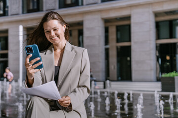 Smiling businesswoman multitasking with a smartphone and documents, standing by a fountain in a sunny city plaza