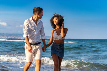 Young interracial couple - walking hand in hand on a sandy beach, enjoying a romantic and joyful...