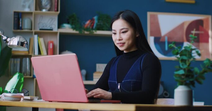 Charming Chinese woman starting up laptop. Beginning typing immediately. Looking at different windows on screen. Smiling with joy. Working remotely. Describing something or recording blog.