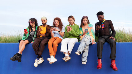 Group of multiracial people sitting outdoors on a blue wall using cell phones together, immersed in their own, watching social media content. Concept of technology addiction generation.