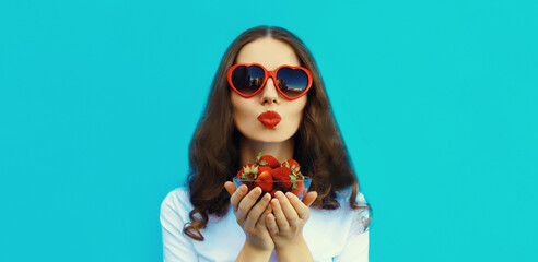 Portrait of beautiful young woman with handful of fresh strawberries blowing kiss on blue background