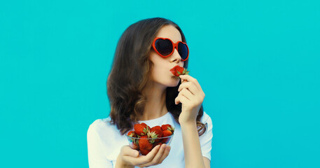 Portrait of beautiful young woman with handful of fresh strawberries blowing kiss on blue background