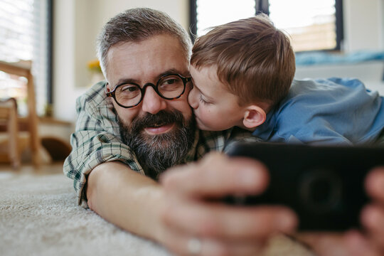 Little boy making selfie on smartphone with father, lying on floor in kids room, making silly faces. Dad explaining technology to son, digital literacy for kids.