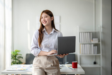 A woman is smiling and holding a laptop in front of a white wall. Concept of productivity and focus, as the woman is likely working on her laptop. The white wall and the organized desk suggest a clean