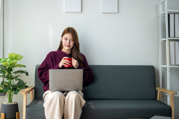 A woman is sitting on a couch with a laptop and a red mug in front of her. She is wearing a purple...
