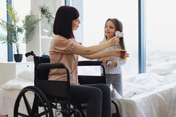 Obraz premium Yong mother wheelchair user brushing her little daughter's hair sitting near big panoramic window in bedroom. Happy Caucasian family having fun on bad on weekend morning.