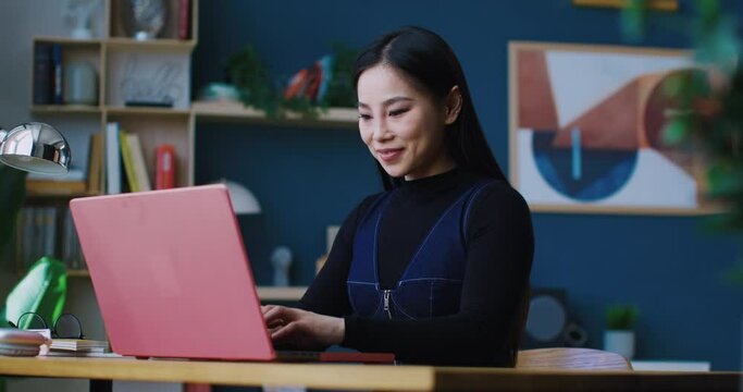 Cute Chinese girl starting up laptop. Sitting in big room full of shelves and paintings. Organized person working from home. Student completing all required work remotely. Concept of job.