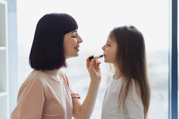 Happy Mother and daughter having fun together at weekend. Little girl doing makeup to mom sitting at bedroom. The daughter paints lips to her beautiful mommy. Happy loving family.