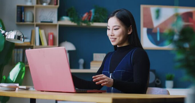 Beautiful Chinese girl getting ready for work remotely. Sitting home at desk while opening up laptop lid. Preparing for productive day. Putting on glasses for better vision. Pressing keys on keyboard.