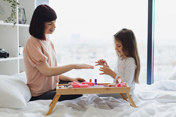 Cute little girl learns to paint her nails by applying polish on mother hands. Happy mother and daughter sitting in bed at home and doing manicure, taking care of their body.