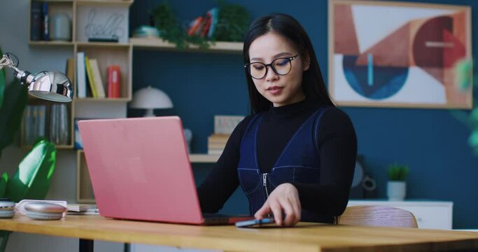 Cute Asian woman sitting in front of laptop. Focused on working remotely while hearing phone ringing. Picking up smartphone. Beginning conversation with co-worker or relative. Distracted girl.