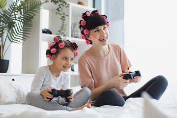 A young woman and her little girl spend their free time together. Happy mother and daughter in curlers sitting on bed at home and playing video games using joysticks.