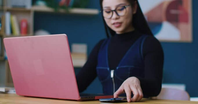 Charming Chinese girl with glasses studying remotely while being distracted by mobile phone ringing. Picking up smartphone. Replying to mobile call. Conversation on digital device.