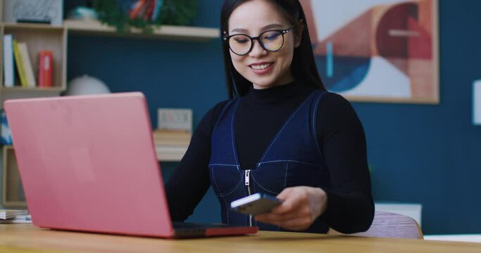 Camera focus on Chinese girls face. Woman with glasses and long straight hair working from home or comfy office. Sitting in front of laptop while talking on phone. Smiling during discussion.