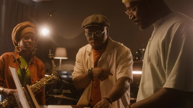 Medium of two African American male musicians standing at electric piano in studio, playing various chords on keyboards, discussing new song, smiling woman in turban, with saxophone listeningMedium of