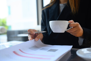Professional businesswoman holding cup of hot coffee and checking financial reports at desk
